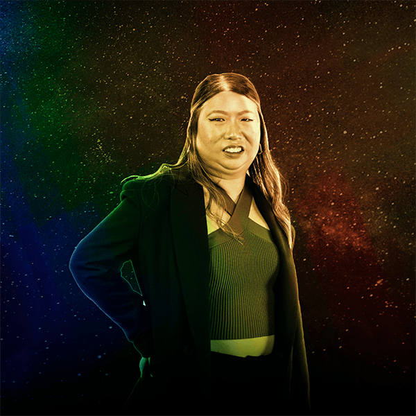 Late-twenties ensemble member, Jan de la Cruz, posing casually with arms on her hip against a star background. The image is treated with a rainbow overlay to match the album artwork. Jan is medium-light skinned, with black hair, wearing a black overcoat, black pants, and brown halter top.