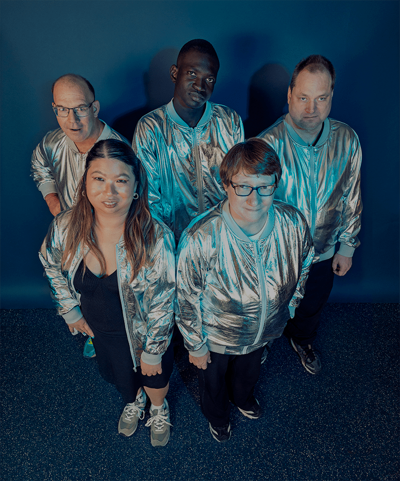 Group image of all five ensemble members wearing silver shiny jackets against a blue photo backdrop. They are standing and looking up at the camera in a trapezoid formation. Jan and Colleen in front, Tony, David and Mark in the back.