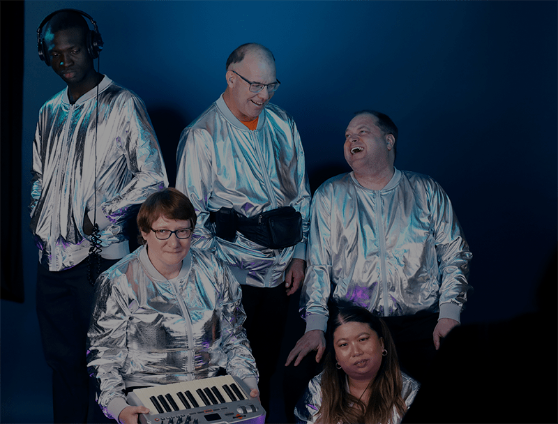 All five music ensemble members looking candid against a blue backdrop during the album artwork photo session. The group are all wearing metallic silver jackets and are casually interacting between posed photos.
