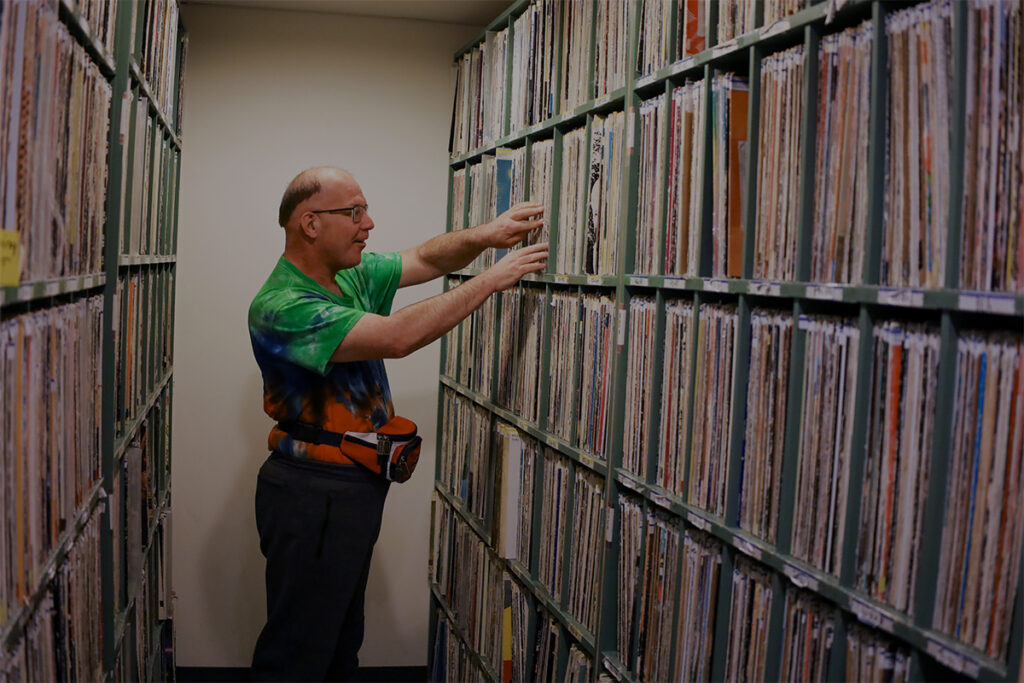 Ensemble member, Tony Goodison, excitedly sorting through a tall and long library of vinyl records with both hands. There is another same-sized and full shelf behind.